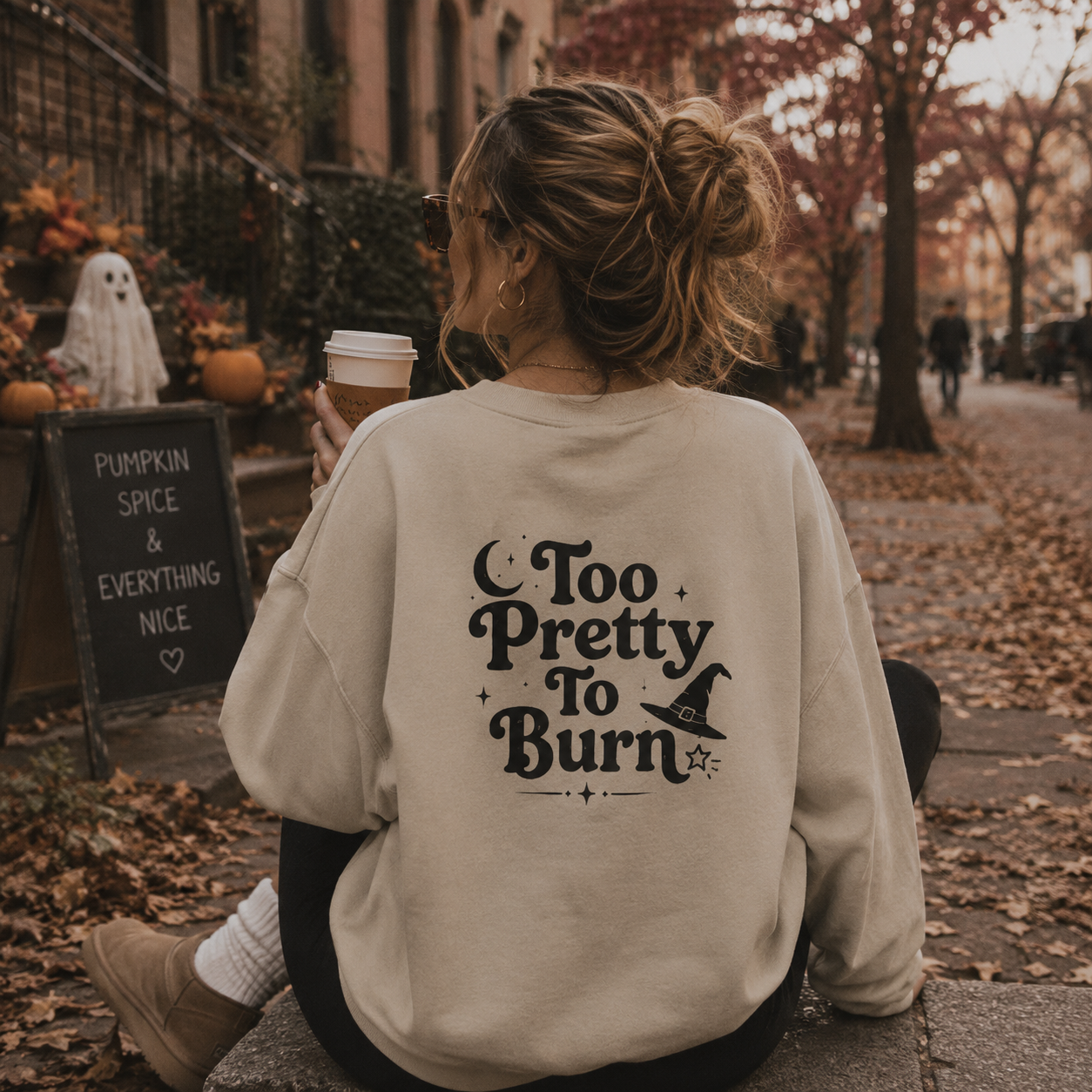 A woman sitting outdoors on a stone path in autumn, viewed from behind, wearing a cream-colored sweatshirt with a black graphic that reads 'Too Pretty To Burn' with a witch hat illustration. She holds a coffee cup, with pumpkins, a ghost decoration, and a cafe chalkboard sign visible in the background.