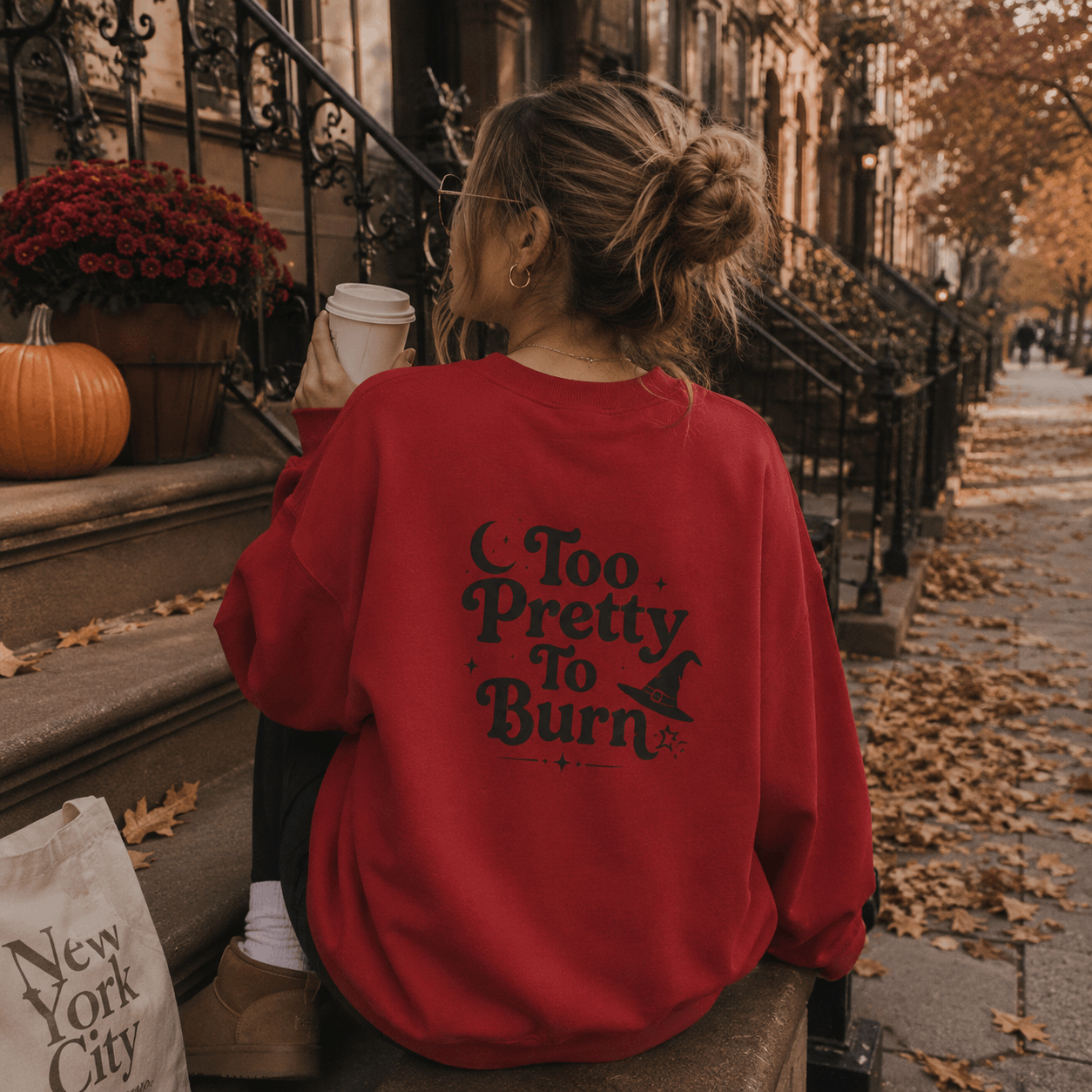A woman sits on a city sidewalk in front of brownstone building steps, seen from behind. She is wearing an oversized red sweatshirt with the phrase 'Too Pretty To Burn' and a witch hat graphic printed in black on the back.
