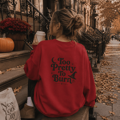 A woman sits on a city sidewalk in front of brownstone building steps, seen from behind. She is wearing an oversized red sweatshirt with the phrase 'Too Pretty To Burn' and a witch hat graphic printed in black on the back.