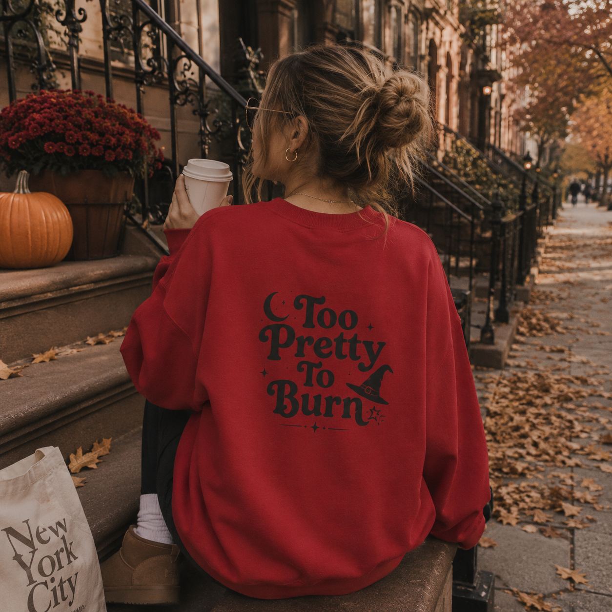 A woman sits on a city sidewalk in front of brownstone building steps, seen from behind. She is wearing an oversized red sweatshirt with the phrase 'Too Pretty To Burn' and a witch hat graphic printed in black on the back.