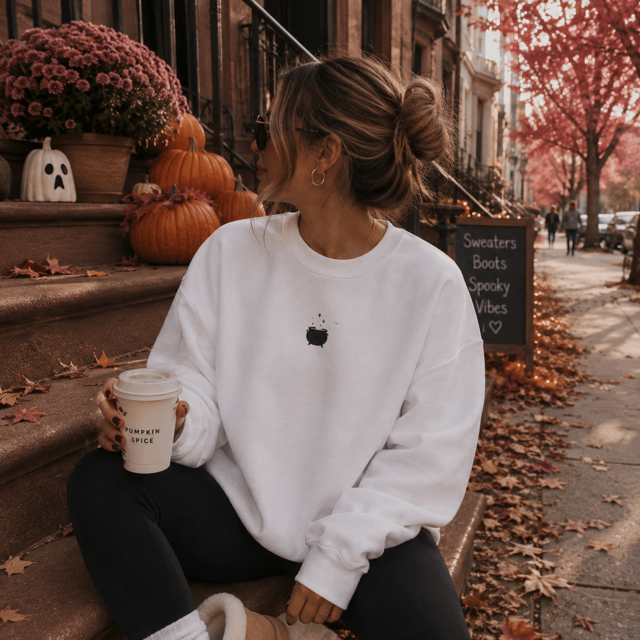 A woman sits on outdoor steps holding a coffee cup, wearing a white crewneck sweatshirt with a small black apple graphic on the chest, black leggings, and beige boots. The background shows autumn street decor with pumpkins, fall leaves, and a chalkboard sign.