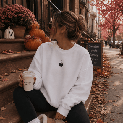 A woman sits on outdoor steps holding a coffee cup, wearing a white crewneck sweatshirt with a small black apple graphic on the chest, black leggings, and beige boots. The background shows autumn street decor with pumpkins, fall leaves, and a chalkboard sign.