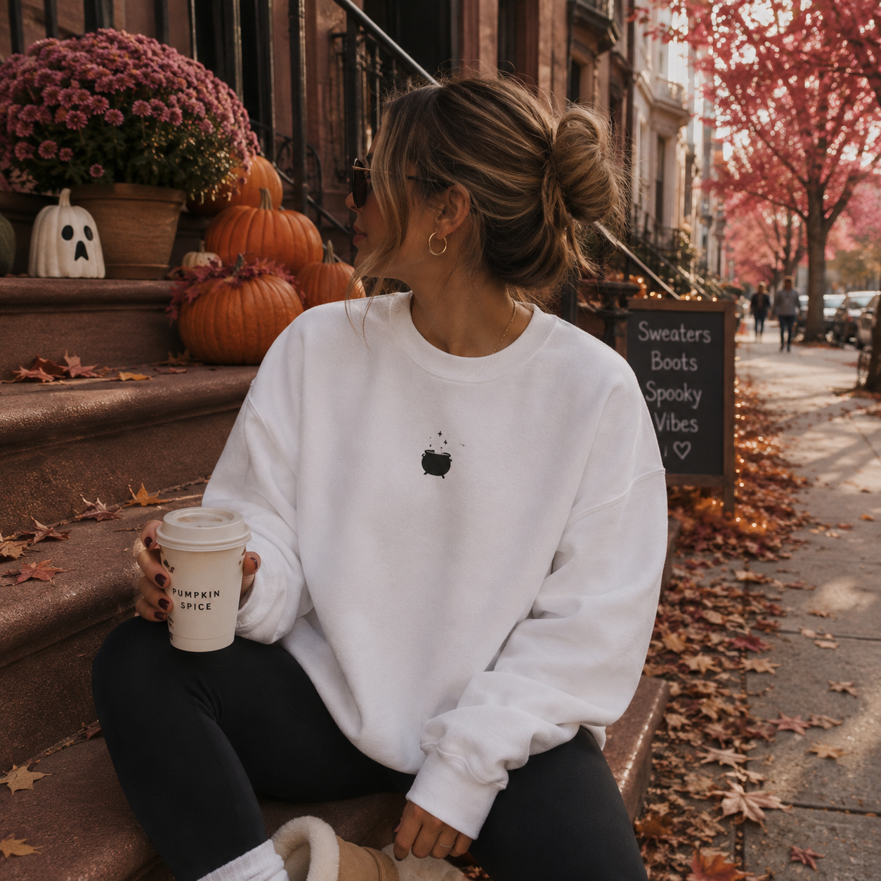 A woman sits on outdoor steps holding a coffee cup, wearing a white crewneck sweatshirt with a small black apple graphic on the chest, black leggings, and beige boots. The background shows autumn street decor with pumpkins, fall leaves, and a chalkboard sign.