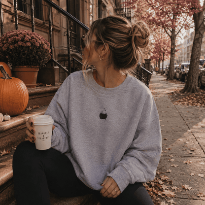 A woman sits on outdoor city steps holding a takeaway coffee cup, wearing an oversized light gray sweatshirt with a small black chest graphic and black pants. Pumpkins, flowers, trees, and a city sidewalk are visible in the background.