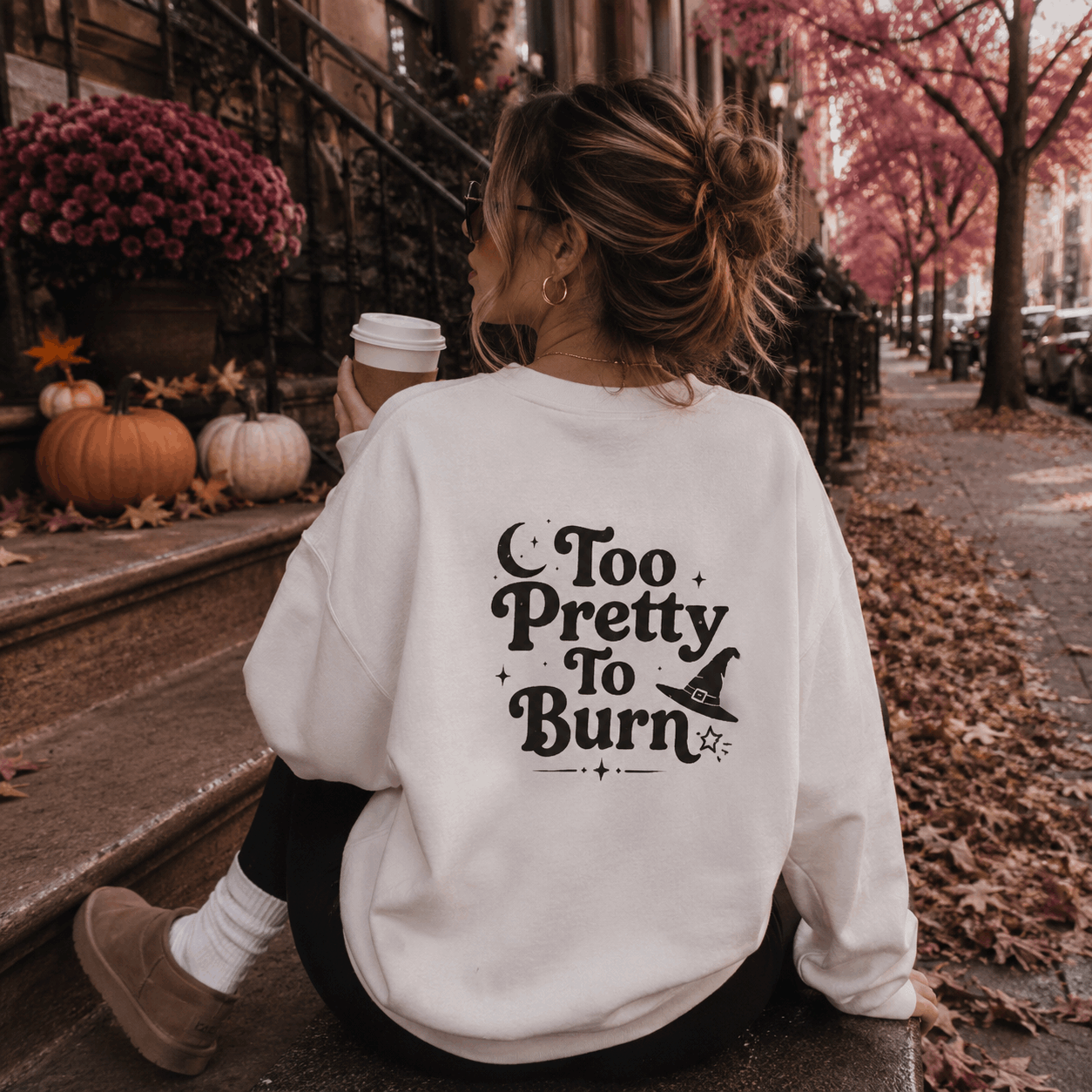 A woman sitting on city steps outdoors, seen from behind, wearing a white sweatshirt with a black decorative text graphic that reads 'Too Pretty To Burn' with a witch hat illustration. The setting includes autumn leaves, pumpkins, flowers, and a coffee cup.