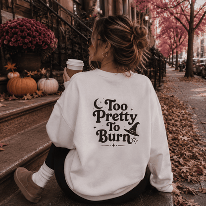 A woman sitting on city steps outdoors, seen from behind, wearing a white sweatshirt with a black decorative text graphic that reads 'Too Pretty To Burn' with a witch hat illustration. The setting includes autumn leaves, pumpkins, flowers, and a coffee cup.