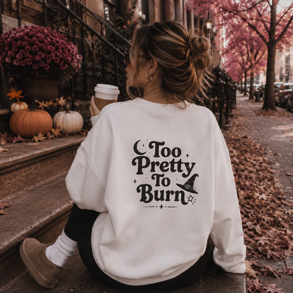 A woman sitting on city steps outdoors, seen from behind, wearing a white sweatshirt with a black decorative text graphic that reads 'Too Pretty To Burn' with a witch hat illustration. The setting includes autumn leaves, pumpkins, flowers, and a coffee cup.