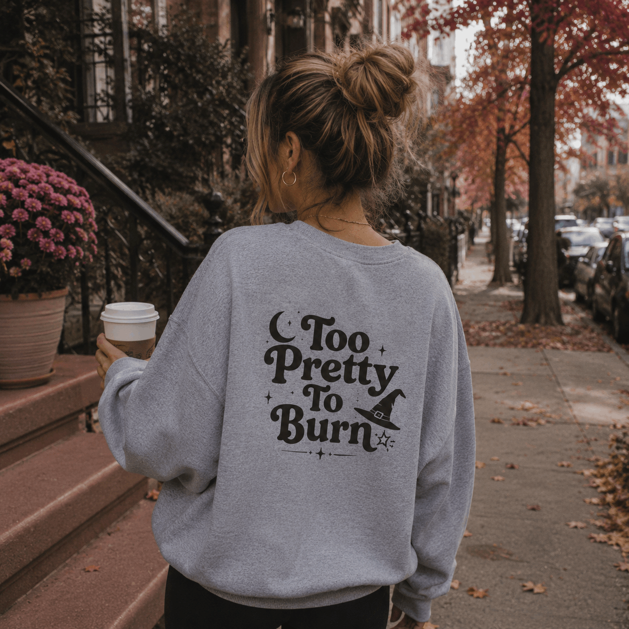 A woman viewed from behind wearing a light gray sweatshirt with a black 'Too Pretty To Burn' witch graphic, standing on a city sidewalk with autumn trees and a coffee cup in hand.