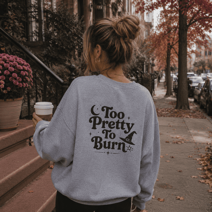 A woman viewed from behind wearing a light gray sweatshirt with a black 'Too Pretty To Burn' witch graphic, standing on a city sidewalk with autumn trees and a coffee cup in hand.