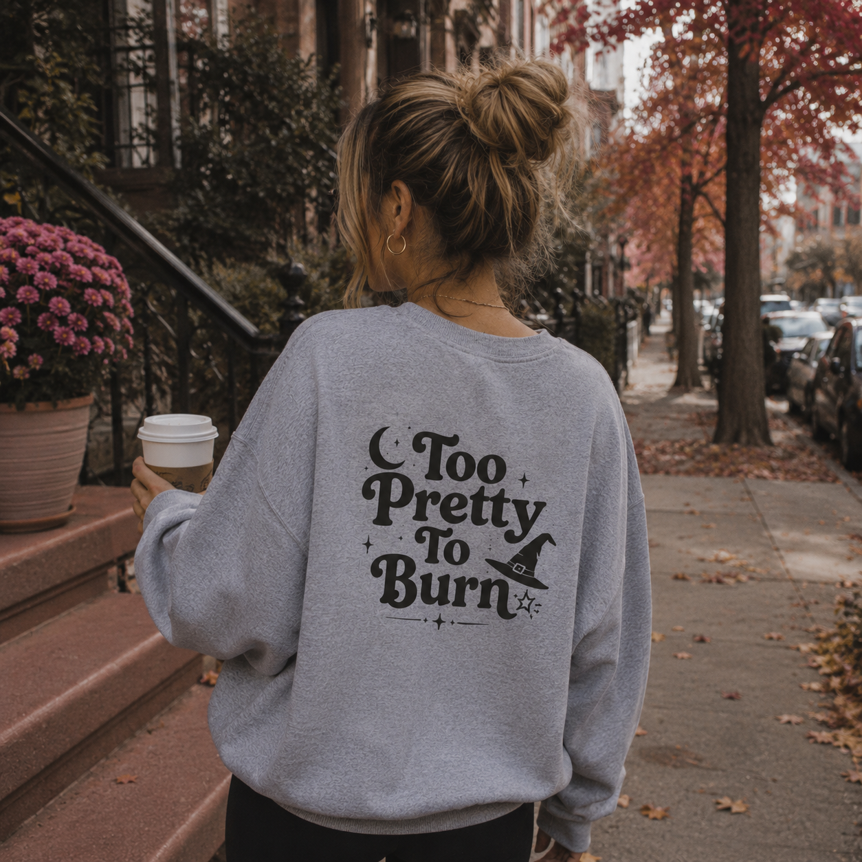 A woman viewed from behind wearing a light gray sweatshirt with a black 'Too Pretty To Burn' witch graphic, standing on a city sidewalk with autumn trees and a coffee cup in hand.