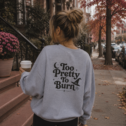 A woman viewed from behind wearing a light gray sweatshirt with a black 'Too Pretty To Burn' witch graphic, standing on a city sidewalk with autumn trees and a coffee cup in hand.
