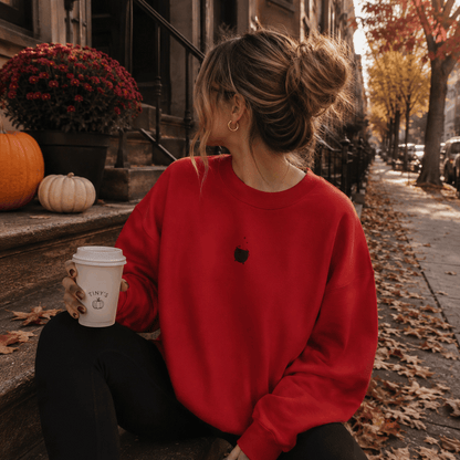 A woman sits on outdoor steps holding a coffee cup, wearing a bright red crewneck sweatshirt with a small black Apple logo on the chest. The background shows a fall street scene with pumpkins, mums, trees with autumn leaves, and a brown building.