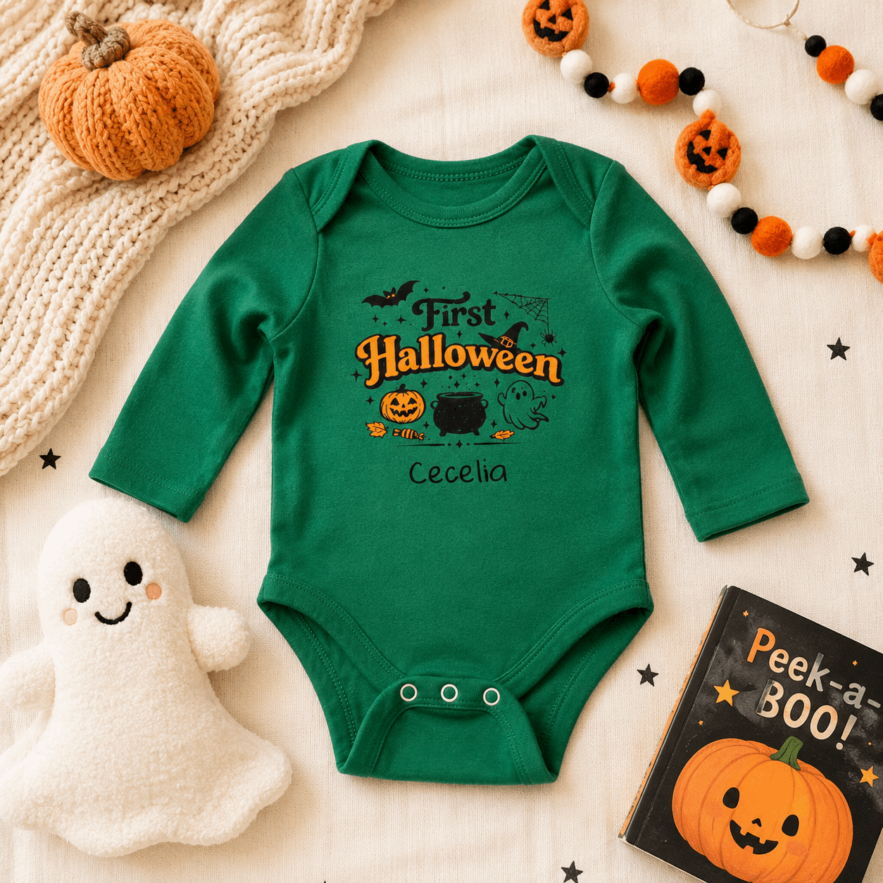 A green long-sleeve baby bodysuit with a 'First Halloween' graphic, laid flat on a cream fabric background with seasonal props including a pumpkin, ghost plush, beads, and a book.
