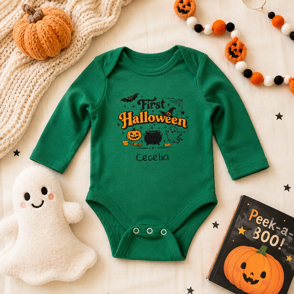A green long-sleeve baby bodysuit with a 'First Halloween' graphic, laid flat on a cream fabric background with seasonal props including a pumpkin, ghost plush, beads, and a book.