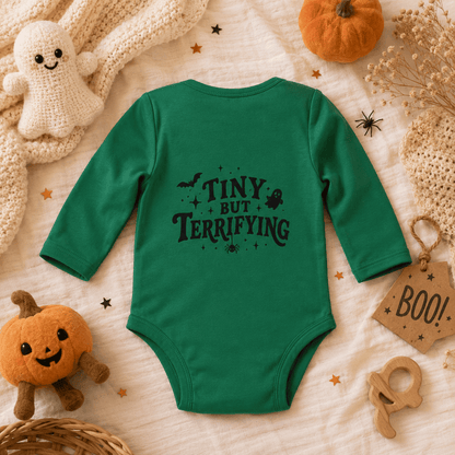 A green long-sleeve baby bodysuit with a black 'TINY BUT TERRIFYING' Halloween graphic, styled in a flat lay with autumn and Halloween props including a small pumpkin, a crochet ghost, and a 'BOO!' tag on a cream fabric background.