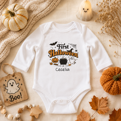 A white long-sleeve baby bodysuit with a 'First Halloween' graphic, styled in a cozy autumn flat-lay with a knit blanket, small pumpkins, dried flowers, a candle, and fall leaves.