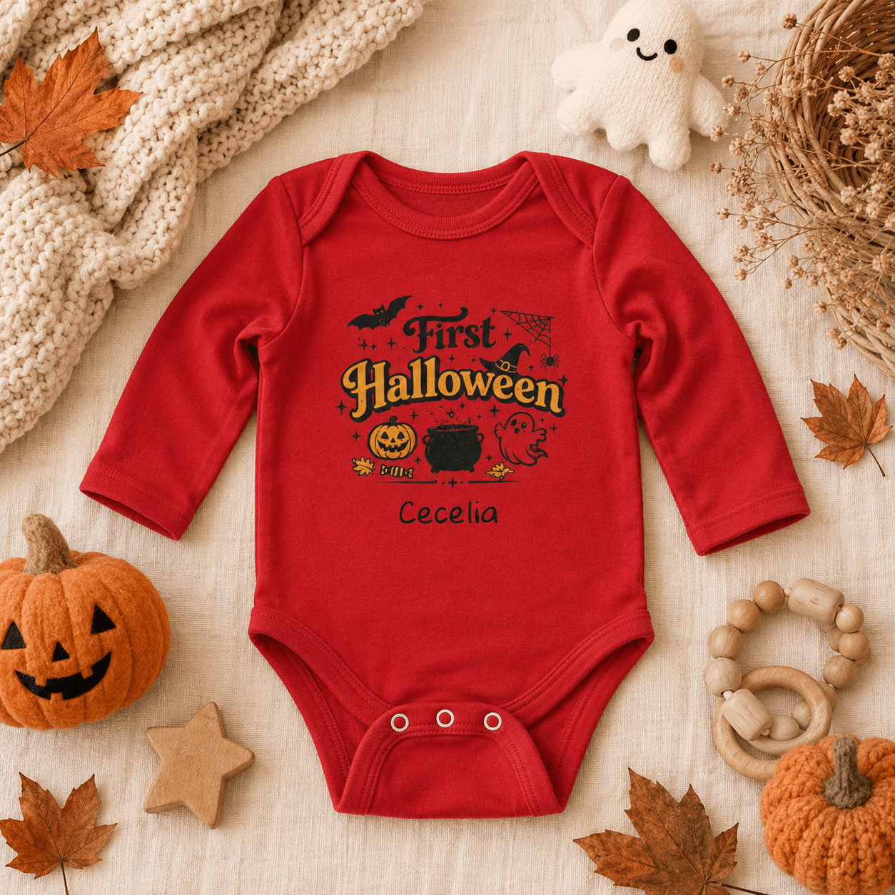 A red long-sleeve baby bodysuit with a 'First Halloween' graphic and the name 'Cecelia' printed on the front, styled in a flat lay with autumn props including a pumpkin, leaves, a knitted blanket, and a plush ghost.