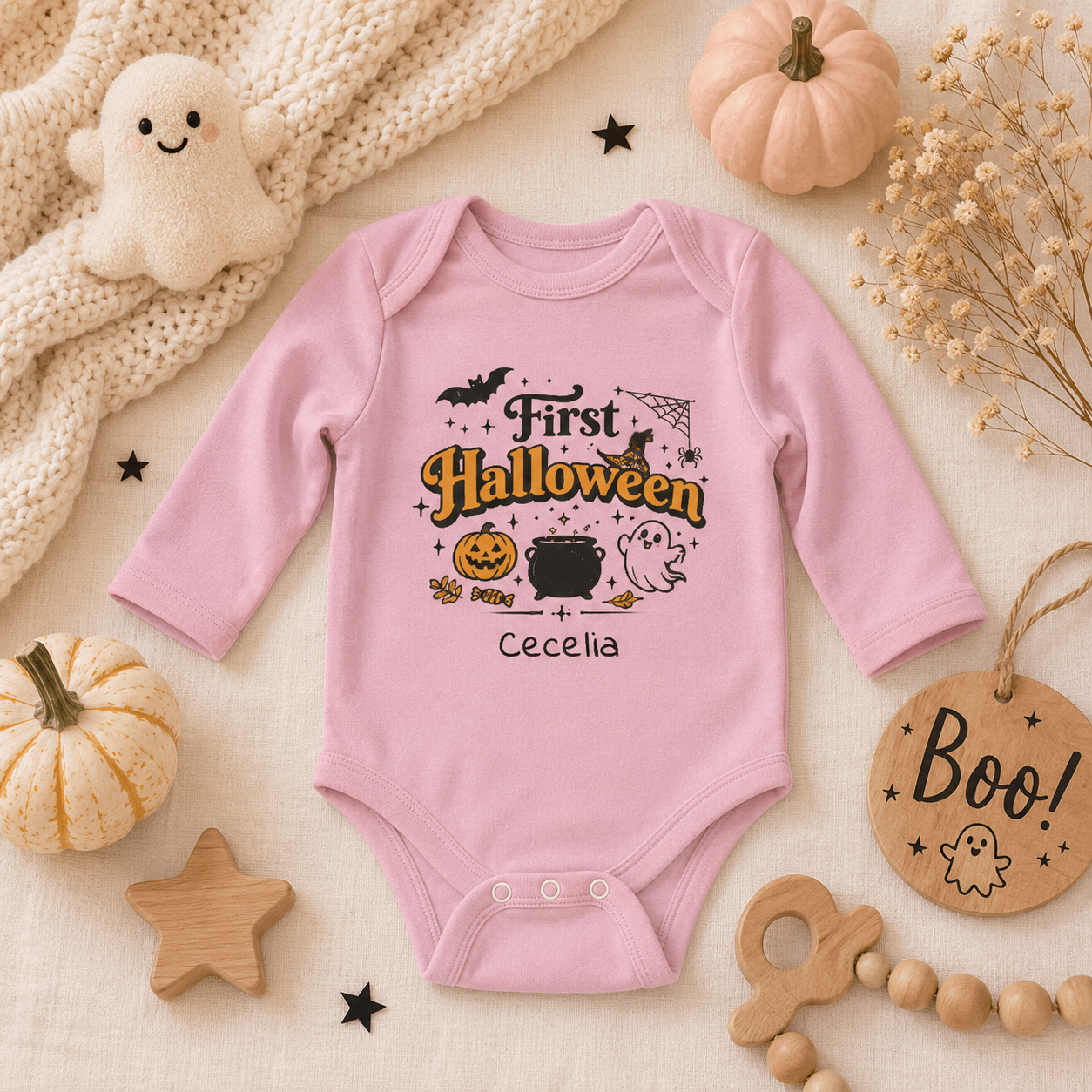 A pink long-sleeve baby bodysuit with a personalized Halloween graphic reading 'First Halloween' and the name 'Cecelia', styled in a flat lay with pumpkins, a plush ghost, wooden stars, beads, and a 'Boo!' tag on a cream fabric background.