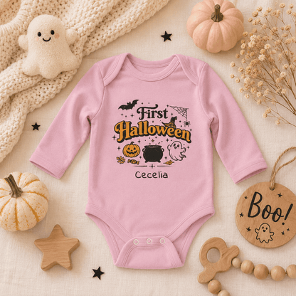 A pink long-sleeve baby bodysuit with a personalized Halloween graphic reading 'First Halloween' and the name 'Cecelia', styled in a flat lay with pumpkins, a plush ghost, wooden stars, beads, and a 'Boo!' tag on a cream fabric background.