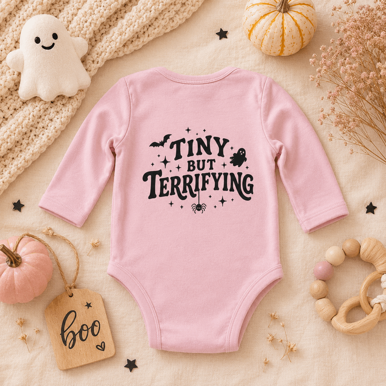 A pink long-sleeve baby bodysuit with a black graphic text reading 'TINY BUT TERRIFYING' surrounded by spooky illustrations, styled in a flat lay with pumpkins, a knit blanket, a wooden tag, and baby accessories.