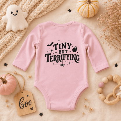 A pink long-sleeve baby bodysuit with a black graphic text reading 'TINY BUT TERRIFYING' surrounded by spooky illustrations, styled in a flat lay with pumpkins, a knit blanket, a wooden tag, and baby accessories.