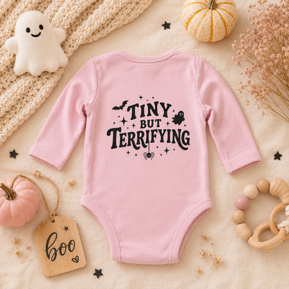A pink long-sleeve baby bodysuit with a black graphic text reading 'TINY BUT TERRIFYING' surrounded by spooky illustrations, styled in a flat lay with pumpkins, a knit blanket, a wooden tag, and baby accessories.
