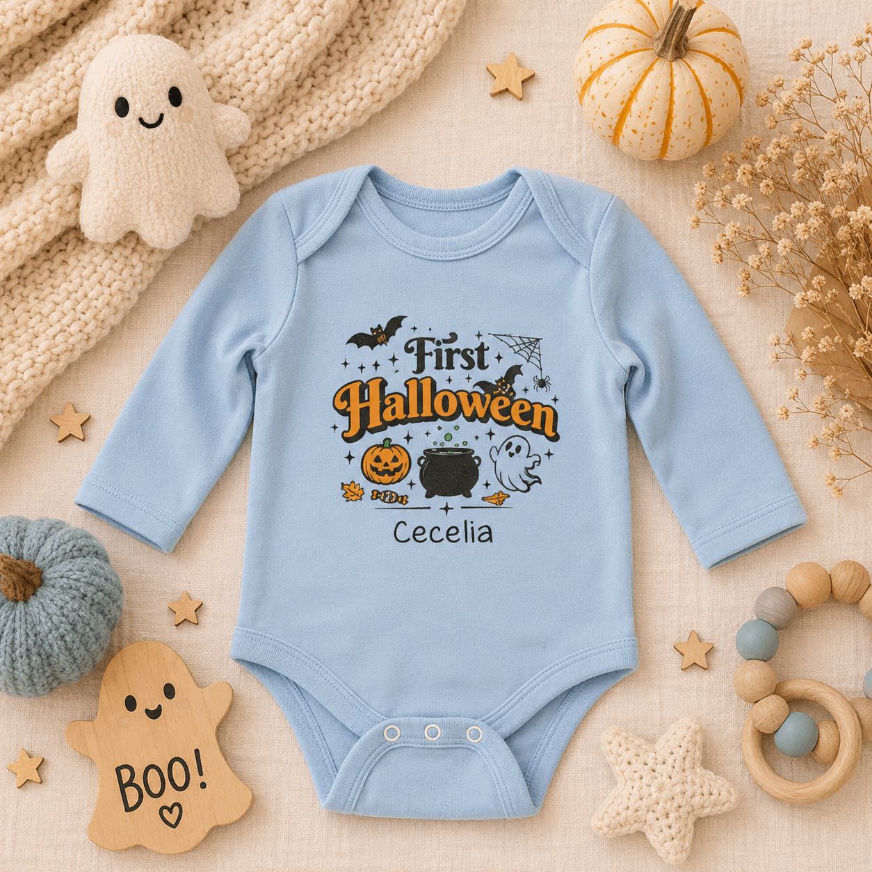 A light blue long-sleeve baby bodysuit with a 'First Halloween' graphic print, styled in a cozy flat-lay arrangement with baby accessories, a plush ghost, and decorative pumpkins on a textured cream fabric background.