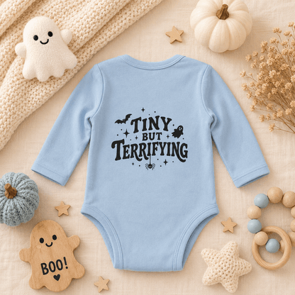 A light blue long-sleeve baby bodysuit with 'TINY BUT TERRIFYING' printed in black on the front, styled in a cozy autumn flat-lay with a knit blanket, small pumpkins, wooden stars, a ghost toy, and baby accessories.