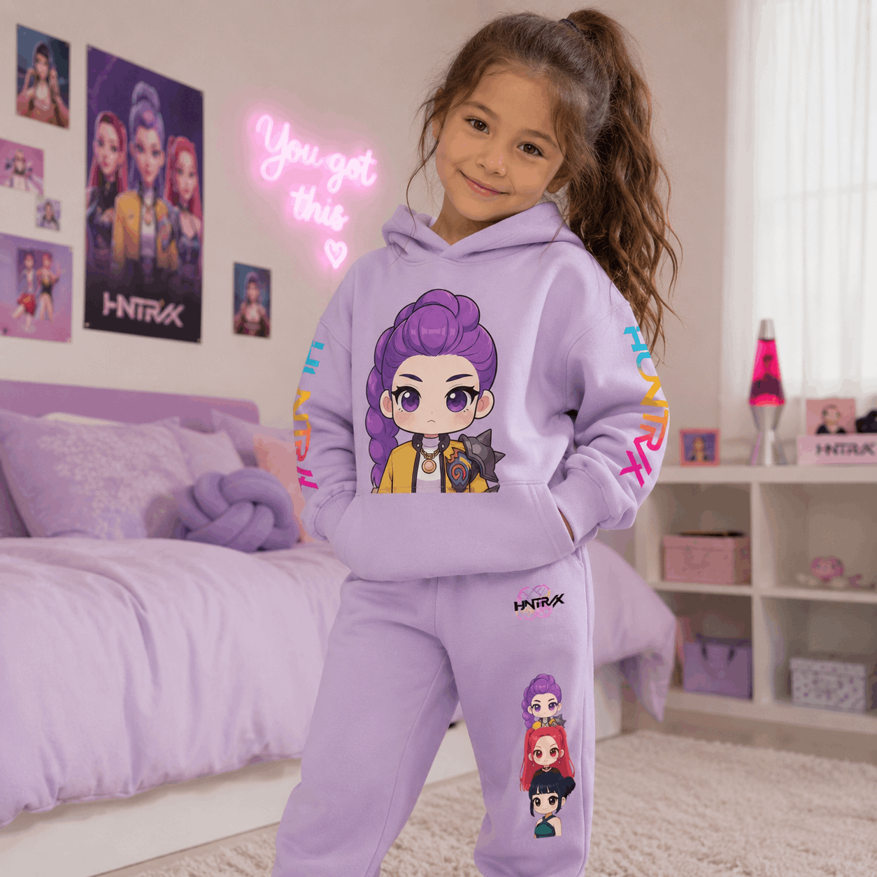 A young girl with her hair in a high ponytail stands in a bedroom wearing a light purple/lavender matching hoodie and sweatpants set with colorful cartoon character prints and HNTRX branding.
