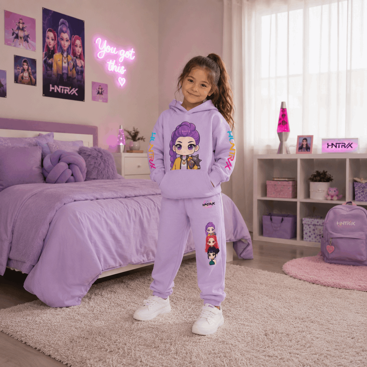 A young girl stands in a bedroom wearing a lavender purple matching hoodie and sweatpants set with colorful cartoon character graphics. The room features a bed, shelves, and a pink neon sign.