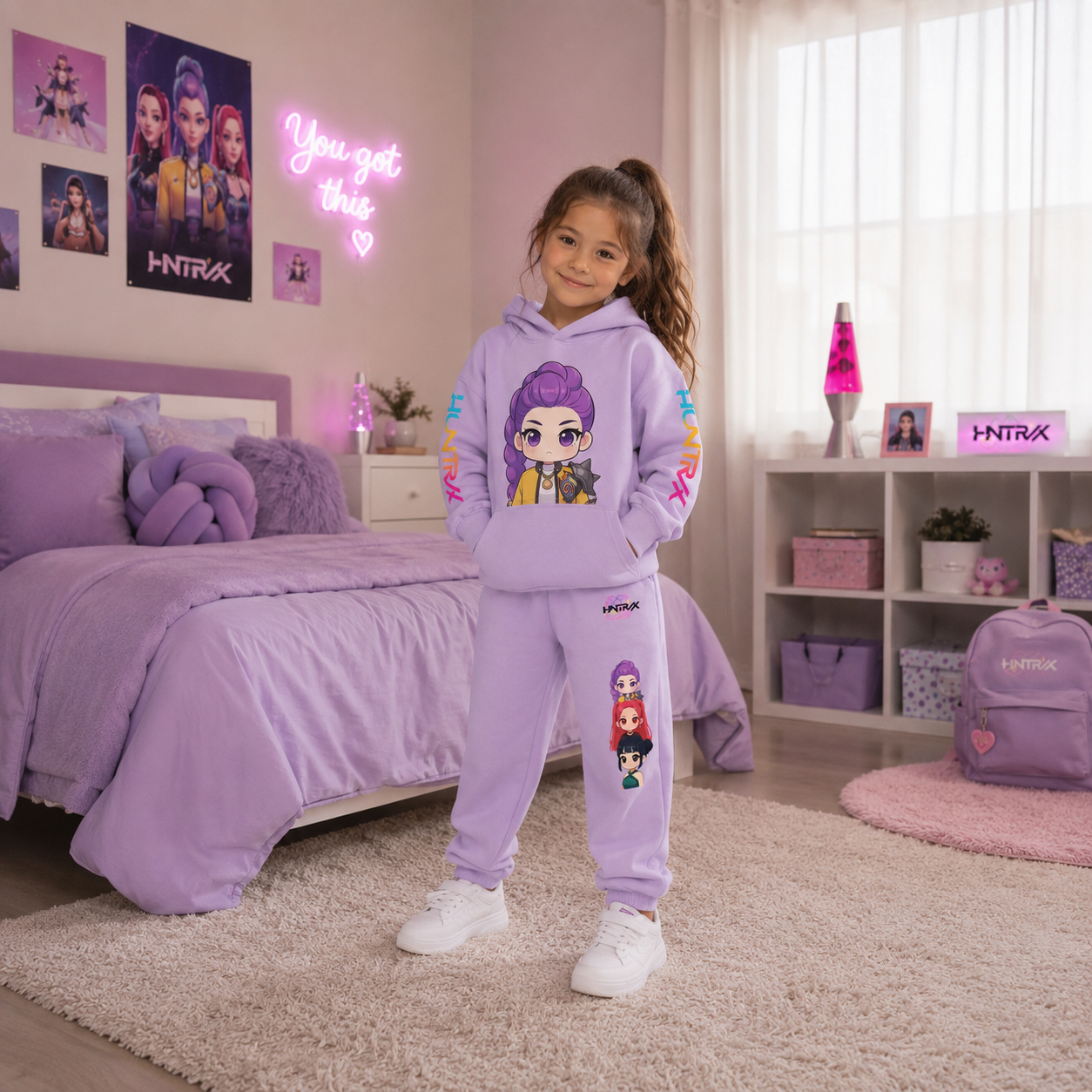 A young girl stands in a bedroom wearing a lavender purple matching hoodie and sweatpants set with colorful cartoon character graphics. The room features a bed, shelves, and a pink neon sign.