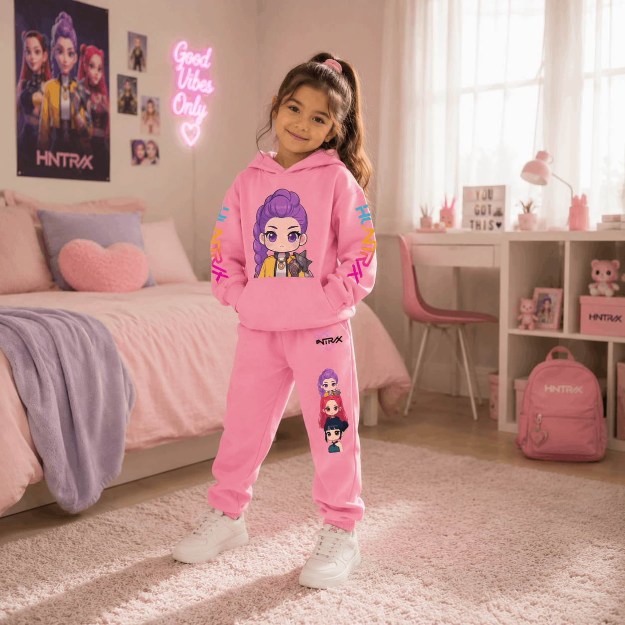A young girl stands in a pink bedroom wearing a matching pink hoodie and sweatpants set with cartoon character graphics. The room has a bed, desk, chair, and pink backpack, with the brand name HINTRAX visible on the wall poster, neon sign, and backpack.