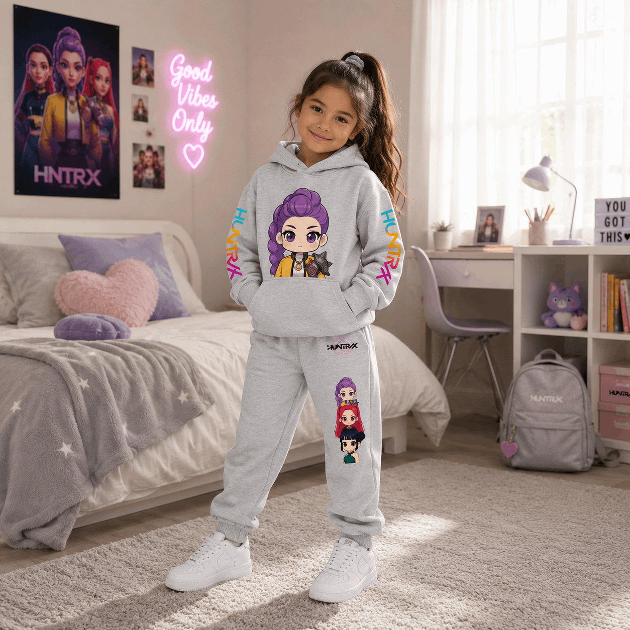 A young girl stands in a bright bedroom wearing a light gray two-piece hoodie and sweatpants set with colorful cartoon character prints and 'SNAPTRX' branding. The room includes a bed, desk, window, and matching branded backpack and wall decor.