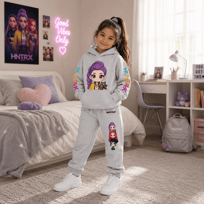 A young girl stands in a bright bedroom wearing a light gray two-piece hoodie and sweatpants set with colorful cartoon character prints and 'SNAPTRX' branding. The room includes a bed, desk, window, and matching branded backpack and wall decor.