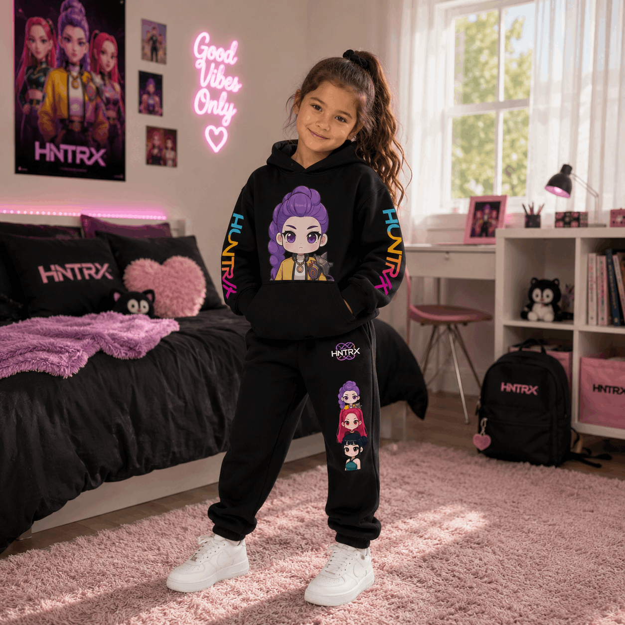 A young girl stands in a pink bedroom wearing a matching black hoodie and sweatpants set with colorful cartoon graphics and the brand name HINTRX. The room features a bed, desk, shelves, and a neon sign.