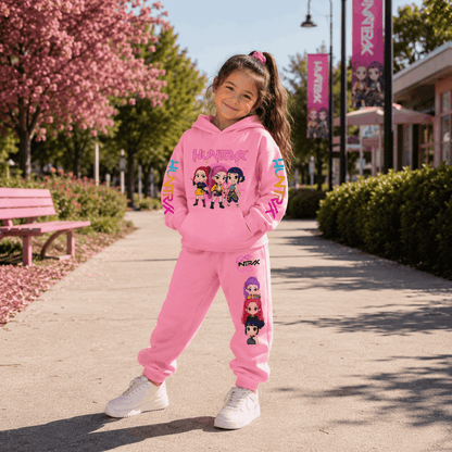A young girl stands on an outdoor paved walkway wearing a pink two-piece tracksuit with cartoon character graphics. The set includes a pullover hoodie and matching jogger pants, with white sneakers.