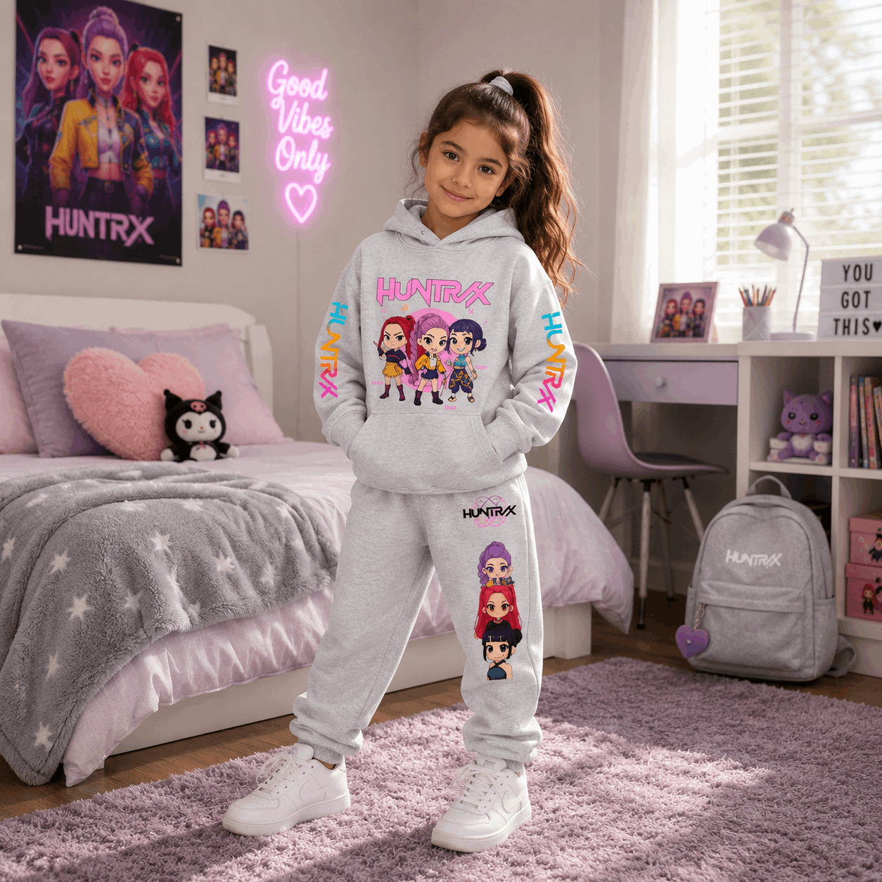 A young girl stands in a bedroom wearing a light gray matching hoodie and sweatpants set with colorful cartoon graphics and the word 'HUNTRX' printed on them. The room has a bed, desk, chair, and a gray backpack with 'HUNTRX' on it.