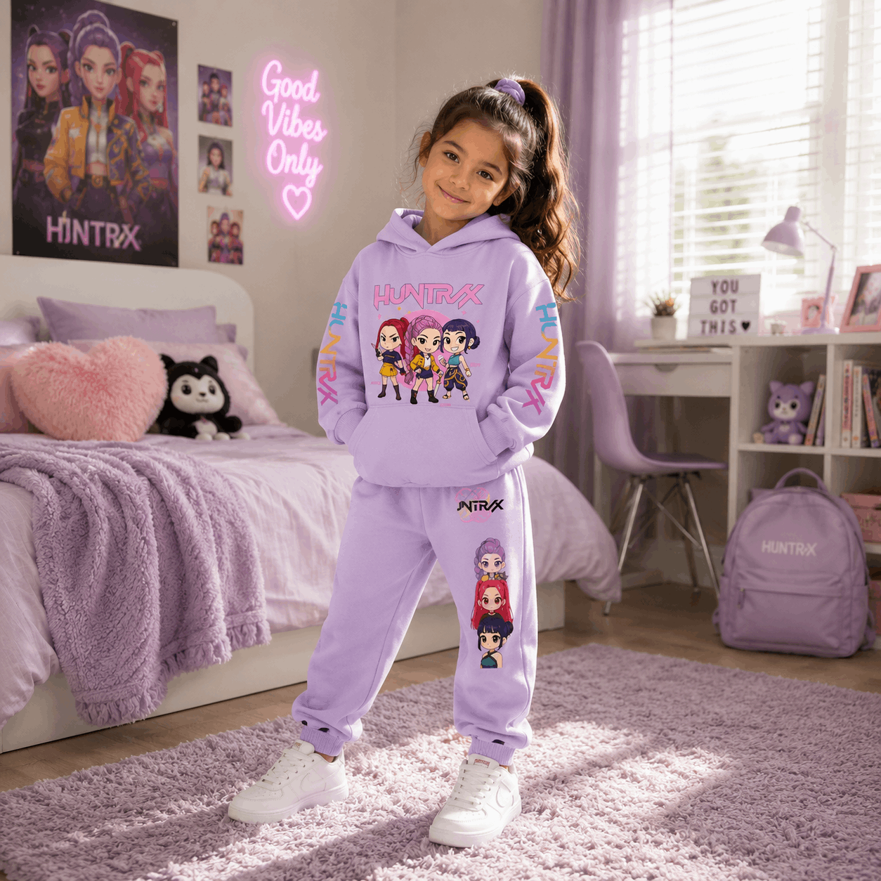 A young girl stands in a bedroom wearing a lavender purple HINTRX matching hoodie and sweatpants set with cartoon character graphics. The room features a bed with purple bedding, a pink heart pillow, a desk, and a purple backpack.