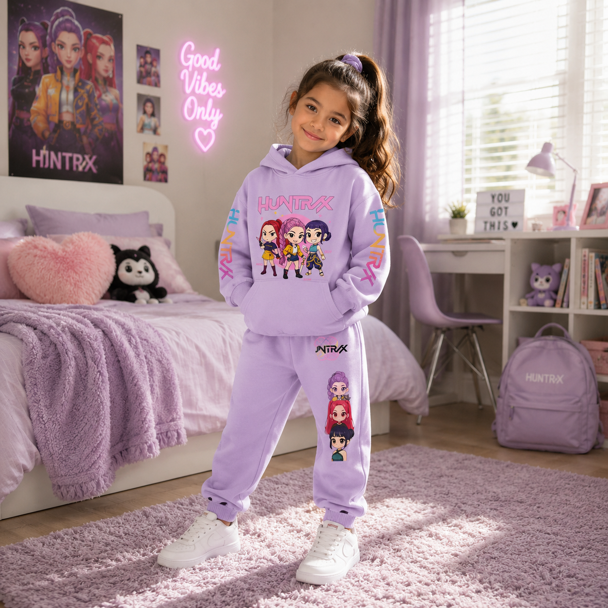 A young girl stands in a bedroom wearing a lavender purple HINTRX matching hoodie and sweatpants set with cartoon character graphics. The room features a bed with purple bedding, a pink heart pillow, a desk, and a purple backpack.