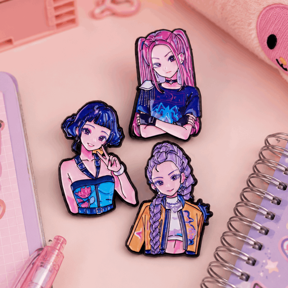 Three colorful anime-style enamel pins with black outlines are arranged on a pink desk surface with stationery props, including a spiral notebook, pen, and plush item.