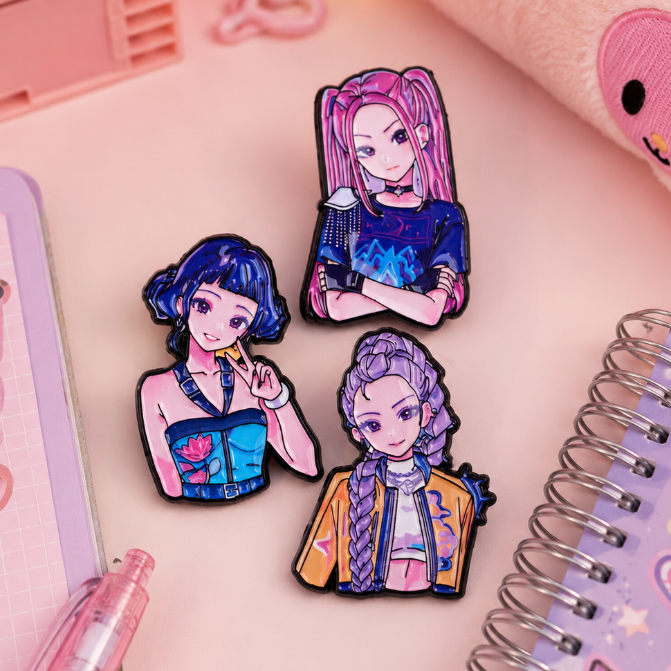 Three colorful anime-style enamel pins with black outlines are arranged on a pink desk surface with stationery props, including a spiral notebook, pen, and plush item.