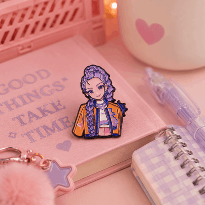 A cute anime-style enamel pin of a girl with purple braided hair, wearing a yellow jacket, displayed on a pink notebook surrounded by stationery props in a pink-themed setting.