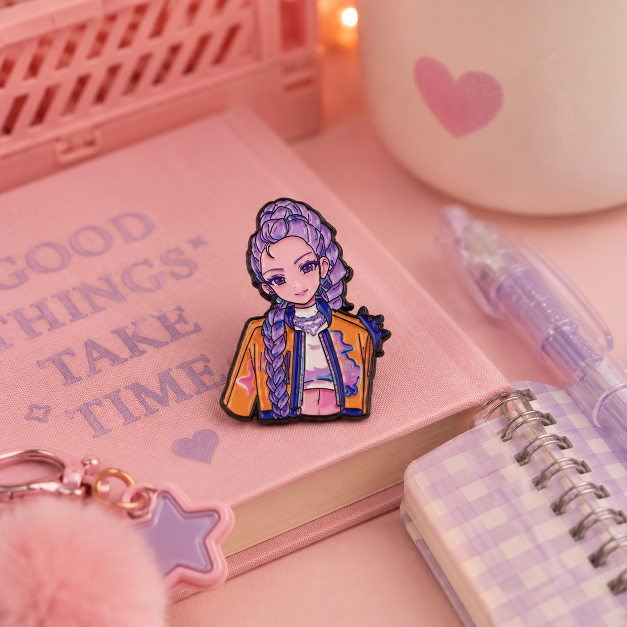 A cute anime-style enamel pin of a girl with purple braided hair, wearing a yellow jacket, displayed on a pink notebook surrounded by stationery props in a pink-themed setting.