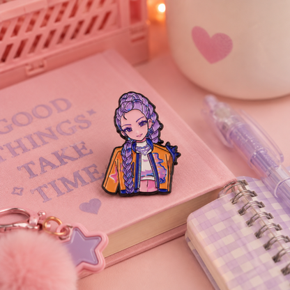A cute anime-style enamel pin of a girl with purple braided hair, wearing a yellow jacket, displayed on a pink notebook surrounded by stationery props in a pink-themed setting.