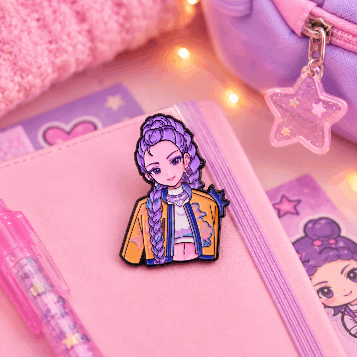 A pink notebook with a purple elastic band, decorated with a cartoon character pin featuring purple braided hair and a yellow jacket. The notebook is styled with a pink pen, a pink textured fabric, and a purple zippered pouch with a star-shaped charm under warm string lights.