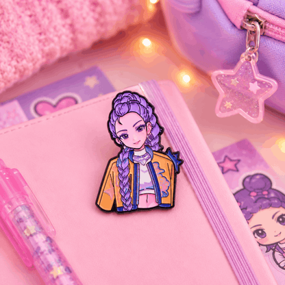 A pink notebook with a purple elastic band, decorated with a cartoon character pin featuring purple braided hair and a yellow jacket. The notebook is styled with a pink pen, a pink textured fabric, and a purple zippered pouch with a star-shaped charm under warm string lights.
