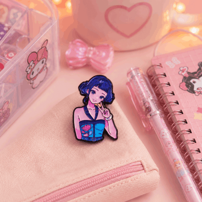 A small enamel pin featuring an anime-style girl with dark blue hair and a pink top, displayed on a light pink canvas zip pouch. The scene is styled with a pink notebook, a pink pen, a pink bow, and a clear organizer box on a soft pink background.