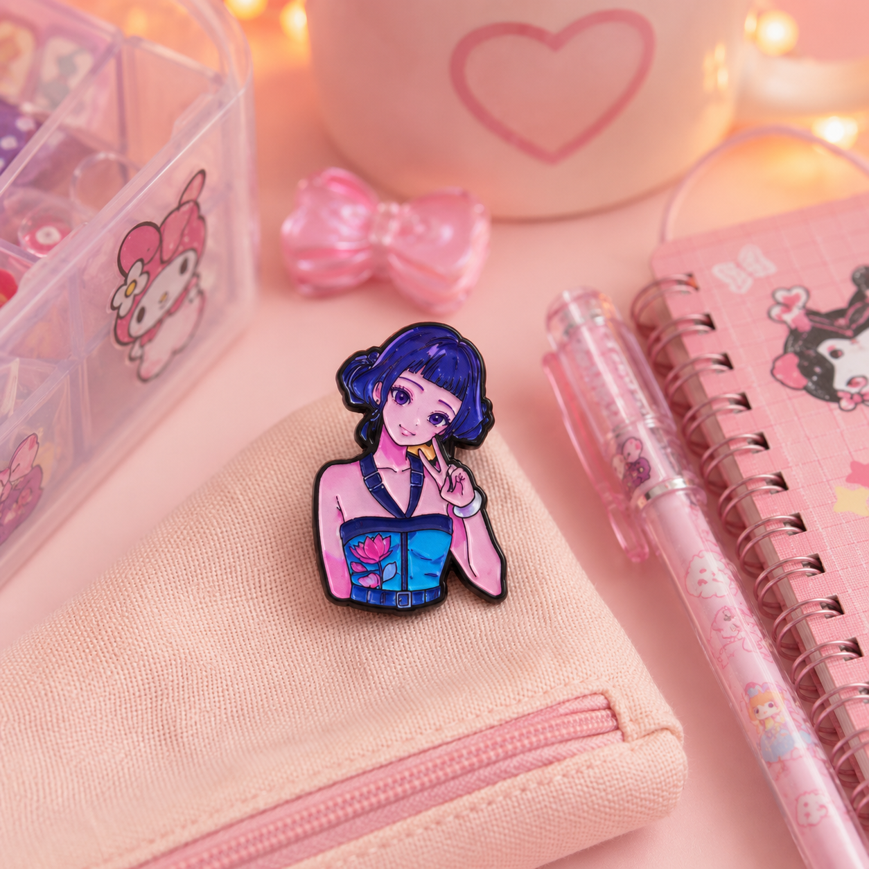 A small enamel pin featuring an anime-style girl with dark blue hair and a pink top, displayed on a light pink canvas zip pouch. The scene is styled with a pink notebook, a pink pen, a pink bow, and a clear organizer box on a soft pink background.