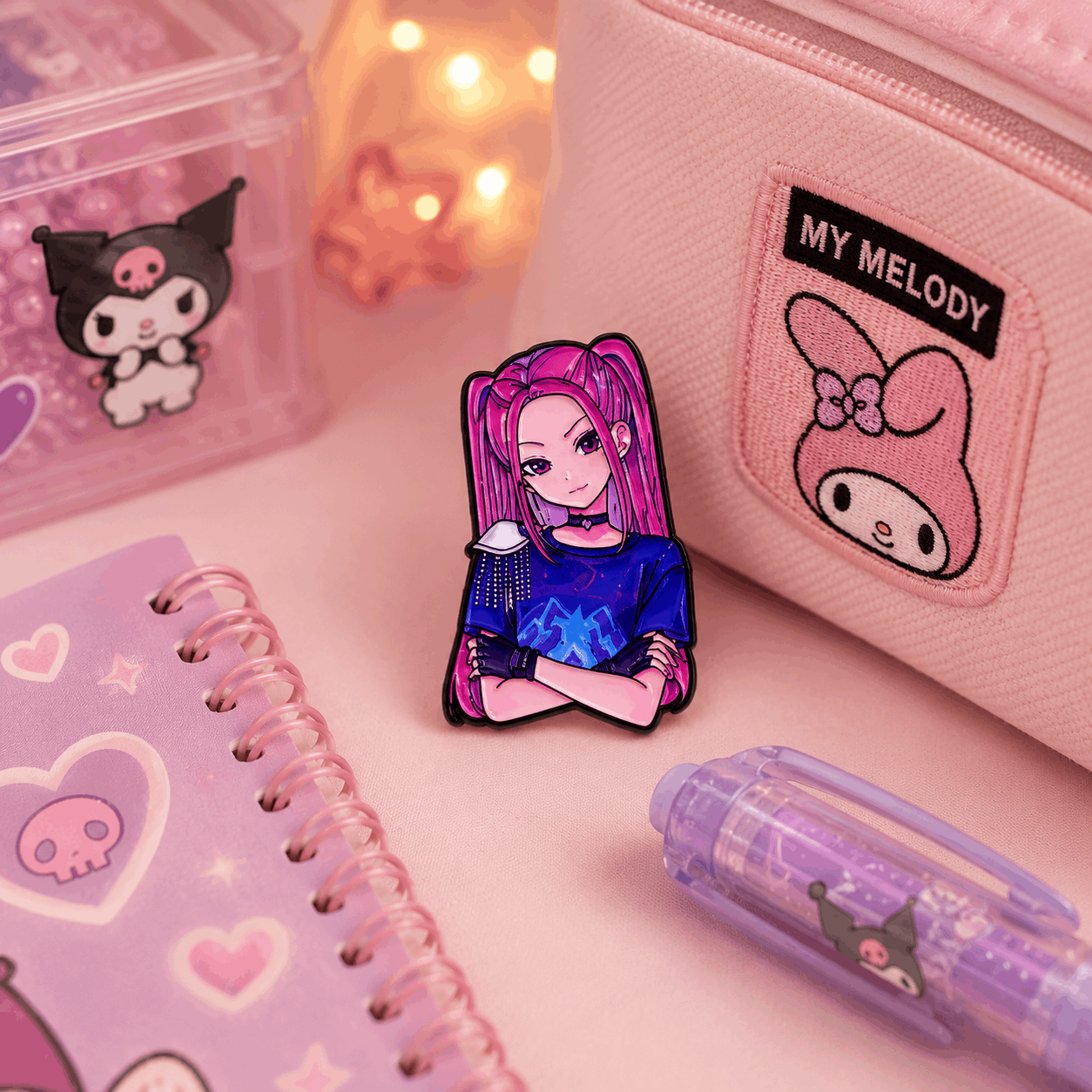A pink and purple enamel pin featuring an anime character with pink hair, styled on a pink desk alongside a pink pencil case, notebook, and pen.