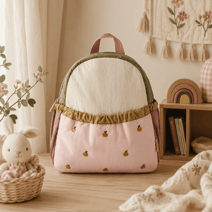 A pastel pink and cream backpack with olive green trim, decorated with small embroidered yellow pineapples, displayed on a wooden table in a cozy, styled room setting.